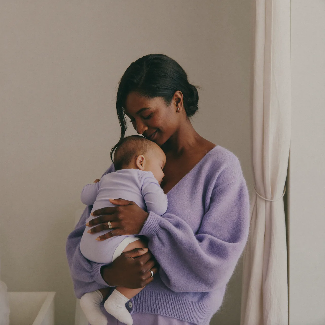 Mãe segurando o bebê adormecido no colo em um quarto tranquilo, em tons de lilás